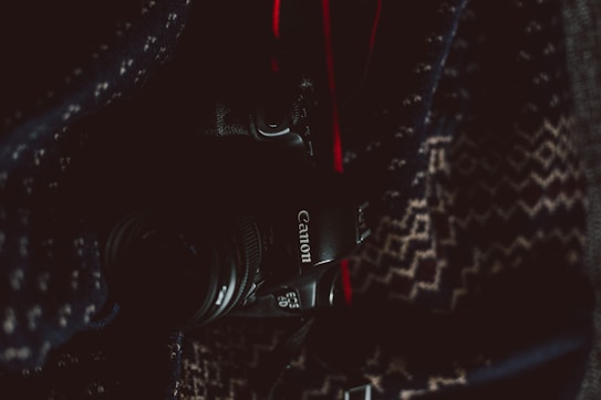 A Canon camera partially visible, surrounded by a patterned fabric in dim lighting. The camera's branding is clearly seen, with parts of the lens and body visible amidst the dark and textured surroundings.