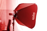 A studio light setup with a red Godox softbox attached to a lighting stand. The scene is lit with a strong red tint, likely from the lighting effect on the softbox. The backdrop is blurred, suggesting a focus on the lighting equipment.