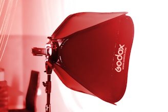 A studio light setup with a red Godox softbox attached to a lighting stand. The scene is lit with a strong red tint, likely from the lighting effect on the softbox. The backdrop is blurred, suggesting a focus on the lighting equipment.