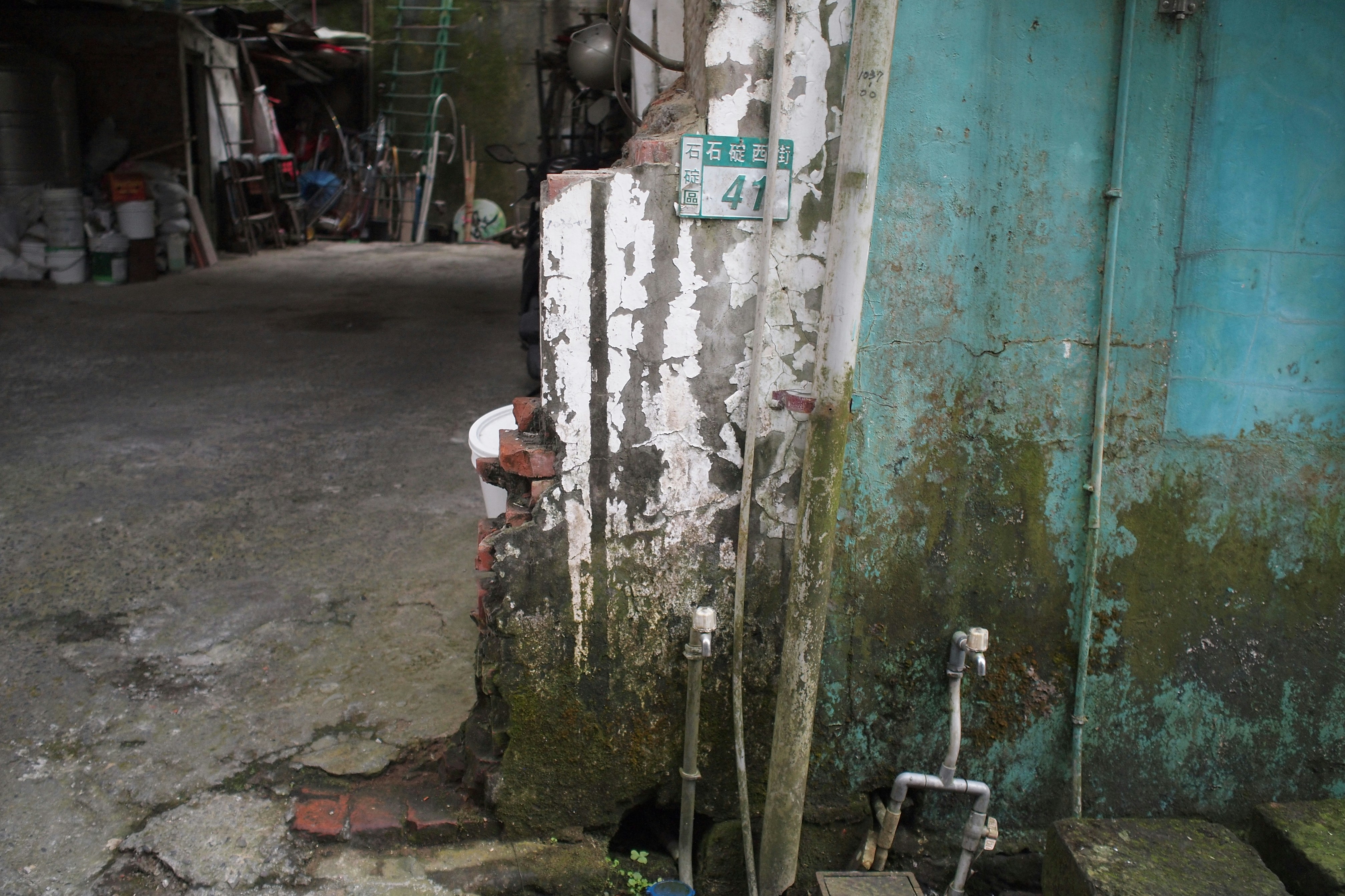An aged, weathered wall with peeling paint and visible water pipes. The wall has a mix of white and teal colors, with signs of moss and grime. Various objects like buckets and tools are scattered around, and there is a ladder leaning against the structure in the background.