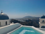 A picturesque view with traditional whitewashed buildings and blue domes beside a turquoise swimming pool. The background features a rugged coastline and a distant mountain under a clear blue sky.