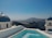 A picturesque view with traditional whitewashed buildings and blue domes beside a turquoise swimming pool. The background features a rugged coastline and a distant mountain under a clear blue sky.