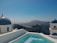 A picturesque view with traditional whitewashed buildings and blue domes beside a turquoise swimming pool. The background features a rugged coastline and a distant mountain under a clear blue sky.