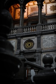 An architectural interior featuring a detailed stone facade with ornate designs. A decorative circular element with intricate patterns is centrally located on the wall, framed by tall pillars and an arched balcony above. Warm lighting from the ceiling creates a contrast with the darker tones of the stone. The foreground includes blurry railings, adding depth to the image.