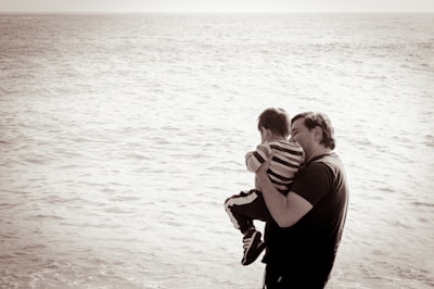 A man is holding a small child in his arms, standing in front of a vast, calm ocean. The image is in sepia tone, and the figures are captured in an intimate moment, facing away towards the water.