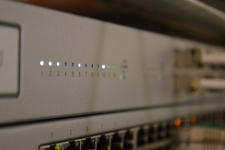 A close-up view of a network switch featuring a row of numbered indicator lights with some illuminated in white and green. The device has multiple Ethernet ports, and connectors are visible in the blurred background.