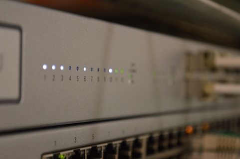 A close-up view of a network switch featuring a row of numbered indicator lights with some illuminated in white and green. The device has multiple Ethernet ports, and connectors are visible in the blurred background.