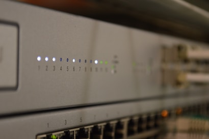 A close-up view of a network switch featuring a row of numbered indicator lights with some illuminated in white and green. The device has multiple Ethernet ports, and connectors are visible in the blurred background.