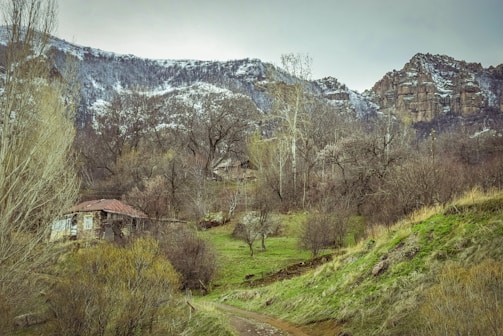 A rustic stone house with a red roof is nestled among lush green fields and leafless trees in a mountainous region. The backdrop features rocky cliffs partially covered with patches of snow, creating a serene and isolated rural setting.