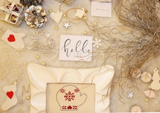 An arrangement of holiday-themed items on a soft, light-colored surface. Included are decorative elements like metallic pine cones, white and silver ornaments, a cross-stitched mitten with red patterns, and handwritten notes with festive messages. Natural twigs add an organic touch.