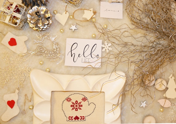 An arrangement of holiday-themed items on a soft, light-colored surface. Included are decorative elements like metallic pine cones, white and silver ornaments, a cross-stitched mitten with red patterns, and handwritten notes with festive messages. Natural twigs add an organic touch.
