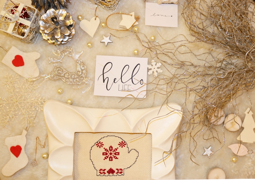 An arrangement of holiday-themed items on a soft, light-colored surface. Included are decorative elements like metallic pine cones, white and silver ornaments, a cross-stitched mitten with red patterns, and handwritten notes with festive messages. Natural twigs add an organic touch.