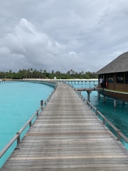 A wooden boardwalk extends over turquoise water, leading to overwater bungalows with thatched roofs. In the background, lush green palm trees border the shoreline under a cloudy sky.