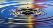 A water droplet is captured in mid-air, creating a series of ripples on a reflective surface. The colorful reflections include shades of blue, yellow, and red, suggesting a vibrant and dynamic environment.
