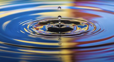 A water droplet is captured in mid-air, creating a series of ripples on a reflective surface. The colorful reflections include shades of blue, yellow, and red, suggesting a vibrant and dynamic environment.