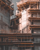 A partially constructed building with concrete structures and wooden scaffolding forming intricate patterns. The architecture is bathed in dim lighting, casting shadows and highlighting the geometric shapes of the construction materials.