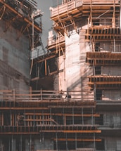 A partially constructed building with concrete structures and wooden scaffolding forming intricate patterns. The architecture is bathed in dim lighting, casting shadows and highlighting the geometric shapes of the construction materials.