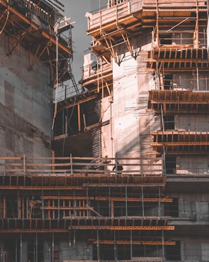 A partially constructed building with concrete structures and wooden scaffolding forming intricate patterns. The architecture is bathed in dim lighting, casting shadows and highlighting the geometric shapes of the construction materials.