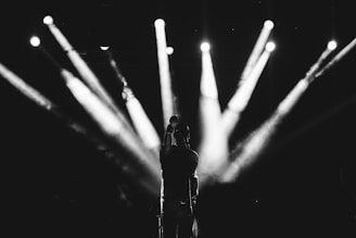 A vocalist stands on stage with bright spotlights radiating from behind, creating a dramatic silhouette. The image is in black and white, emphasizing the contrast between light and shadow.