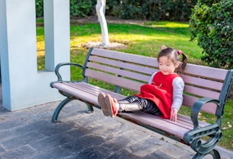 A young child wearing a red dress and black leggings is sitting comfortably on a wooden bench in a park area. The child is leaning back, with legs stretched out and feet crossed. The surroundings include green grass, trees, and a white column structure nearby.