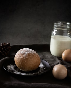 A rustic scene featuring a round, flour-dusted bread roll resting on a dark plate. Nearby, two brown eggs and an open glass jar filled with milk are positioned on a textured, dark cloth. A pinecone rests to the side, adding an organic touch.