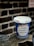 A blue and white disposable coffee cup with the phrase 'It's Our Pleasure to Serve You' printed on the front is placed on a textured surface, possibly metal, with a blurred brick wall in the background.
