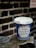 A blue and white disposable coffee cup with the phrase 'It's Our Pleasure to Serve You' printed on the front is placed on a textured surface, possibly metal, with a blurred brick wall in the background.