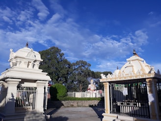 The image features a Hindu temple complex with intricately designed structures. Two small shrines, with detailed carvings and spires, stand prominently. In the background, there is a large statue, likely of a deity, surrounded by greenery. The sky is bright blue with scattered white clouds.
