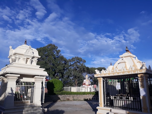 The image features a Hindu temple complex with intricately designed structures. Two small shrines, with detailed carvings and spires, stand prominently. In the background, there is a large statue, likely of a deity, surrounded by greenery. The sky is bright blue with scattered white clouds.