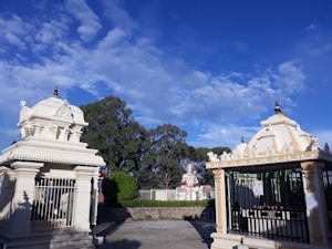 The image features a Hindu temple complex with intricately designed structures. Two small shrines, with detailed carvings and spires, stand prominently. In the background, there is a large statue, likely of a deity, surrounded by greenery. The sky is bright blue with scattered white clouds.