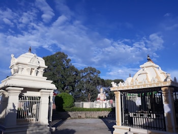 The image features a Hindu temple complex with intricately designed structures. Two small shrines, with detailed carvings and spires, stand prominently. In the background, there is a large statue, likely of a deity, surrounded by greenery. The sky is bright blue with scattered white clouds.