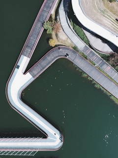 An aerial view of a large body of water intersected by a series of angular walkways. There are clear pathways with wooden boards and a white railing on either side. Green trees and shrubs are visible near the top. Small objects, possibly fish or debris, are seen floating in the water.
