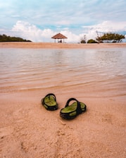Two sandals rest on the sandy shore of a beach. In the background, a small thatched-roof shelter sits on the sand with two people underneath, surrounded by a few trees. The scene includes calm, rippling water in the foreground and a sky filled with scattered clouds.