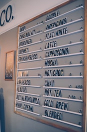 A coffee shop menu board displays various drink options such as drip coffee, cafe au lait, pour over, and espresso. The menu is organized with white letter tiles on a grey background, and prices are listed next to each beverage. There is a wooden frame around the menu board, and a small decorative chalkboard is visible on the left side.