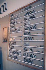 A coffee shop menu board displays various drink options such as drip coffee, cafe au lait, pour over, and espresso. The menu is organized with white letter tiles on a grey background, and prices are listed next to each beverage. There is a wooden frame around the menu board, and a small decorative chalkboard is visible on the left side.