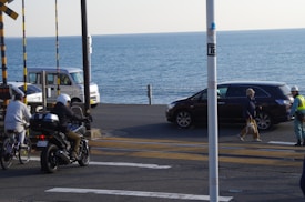 A coastal road features several vehicles and people. A motorcycle and a cyclist are moving along the road, while a dark car and a white van are parked nearby. The background reveals a clear blue ocean, adding a serene atmosphere. Two people are walking, one carrying a plastic bag, and a worker in a safety vest seems to be on duty.