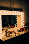 A vanity desk with a mirror framed by warm lights, styled with makeup and accessories.