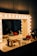 A well-lit vanity mirror surrounded by light bulbs is positioned on a makeup table. The table is cluttered with various cosmetic products, including brushes, palettes, bottles, and other makeup tools. In the reflection of the mirror, a dimly lit background with vague silhouettes of people can be seen.