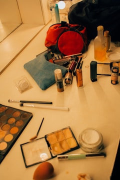 A vanity table is filled with various makeup items, including brushes, palettes, lipsticks, and foundations. A red makeup bag is prominently placed among the products. Other visible items include a blue pouch, skincare bottles, and a mirror in the background. The scene is set in warm lighting, creating a cozy atmosphere.