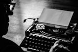 A silhouette of a person wearing glasses is focused on using a vintage typewriter, with a blank sheet of paper visible. The scene is highlighted in black and white shades, emphasizing the nostalgic and timeless feel of the act of typing.