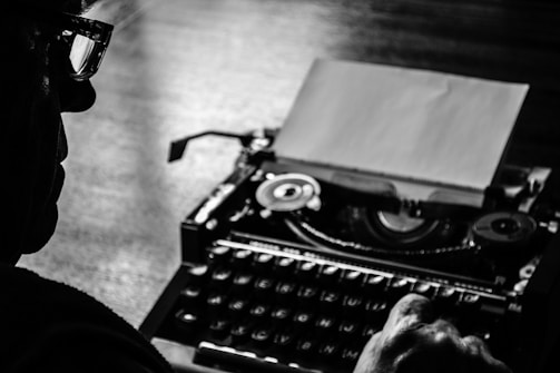 A silhouette of a person wearing glasses is focused on using a vintage typewriter, with a blank sheet of paper visible. The scene is highlighted in black and white shades, emphasizing the nostalgic and timeless feel of the act of typing.