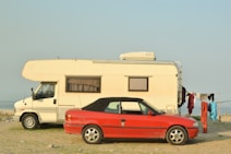 A white camper van and a red convertible car are parked on a grassy shore next to a body of water. Clothes hang to dry on a line beside the vehicles.
