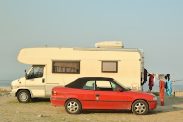 A white camper van and a red convertible car are parked on a grassy shore next to a body of water. Clothes hang to dry on a line beside the vehicles.