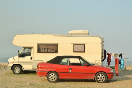 A white camper van and a red convertible car are parked on a grassy shore next to a body of water. Clothes hang to dry on a line beside the vehicles.