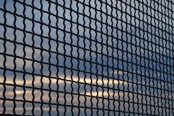 A chain-link fence creates a textured pattern against a backdrop of a sky with clouds and sunlight breaking through. The lines of the fence run diagonally across the image, dividing the view of the sky into small, repeating sections.