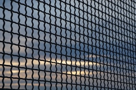 A chain-link fence creates a textured pattern against a backdrop of a sky with clouds and sunlight breaking through. The lines of the fence run diagonally across the image, dividing the view of the sky into small, repeating sections.