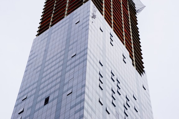 A tall skyscraper with a modern glass facade displaying a pattern of rectangular windows. The upper section is under construction, featuring open spaces with visible red beams.