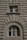 Two windows are set into a textured stone facade. The upper window is rectangular, while the lower one is arched, both with beige frames contrasting against the intricately carved surface that features protruding geometric patterns.