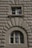 Two windows are set into a textured stone facade. The upper window is rectangular, while the lower one is arched, both with beige frames contrasting against the intricately carved surface that features protruding geometric patterns.