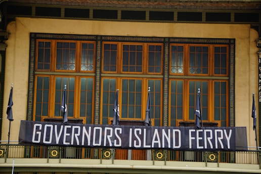 A large banner with the words 'Governors Island Ferry' is prominently displayed across a classic architectural facade featuring large wooden-framed windows. Several black flags are lined up along the structure, contributing to a formal and structured atmosphere.